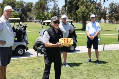 A golfer holding a dice game box at a hole contest, with golf carts behind